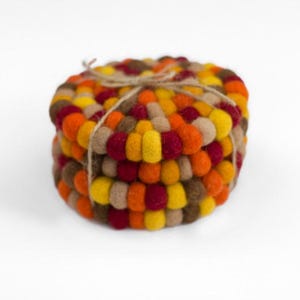 May include: A stack of circular felt ball coasters tied together with a thin brown twine string. The coasters are constructed from small, round, felted wool spheres in a vibrant autumn color palette of deep red, bright yellow, orange, and tan.