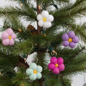 May include: A collection of felt flower ornaments in various colors, including pink, white, purple, green, and magenta, with yellow centers. The ornaments are hanging on a Christmas tree with green needles and small lights.