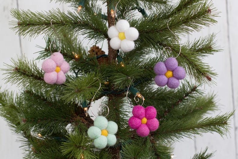 May include: A collection of felt flower ornaments in various colors, including pink, white, purple, green, and magenta, with yellow centers. The ornaments are hanging on a Christmas tree with green needles and small lights.