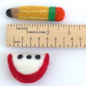 May include: A felt apple slice and a felt pencil, both about 2 inches long, are placed next to a wooden ruler with inch and centimeter markings. The apple slice is red with a white center and three black seeds. The pencil is yellow with a black eraser and green and orange stripes at the end.