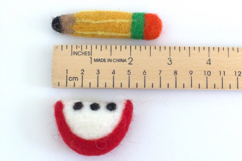 May include: A felt apple slice and a felt pencil, both about 2 inches long, are placed next to a wooden ruler with inch and centimeter markings. The apple slice is red with a white center and three black seeds. The pencil is yellow with a black eraser and green and orange stripes at the end.