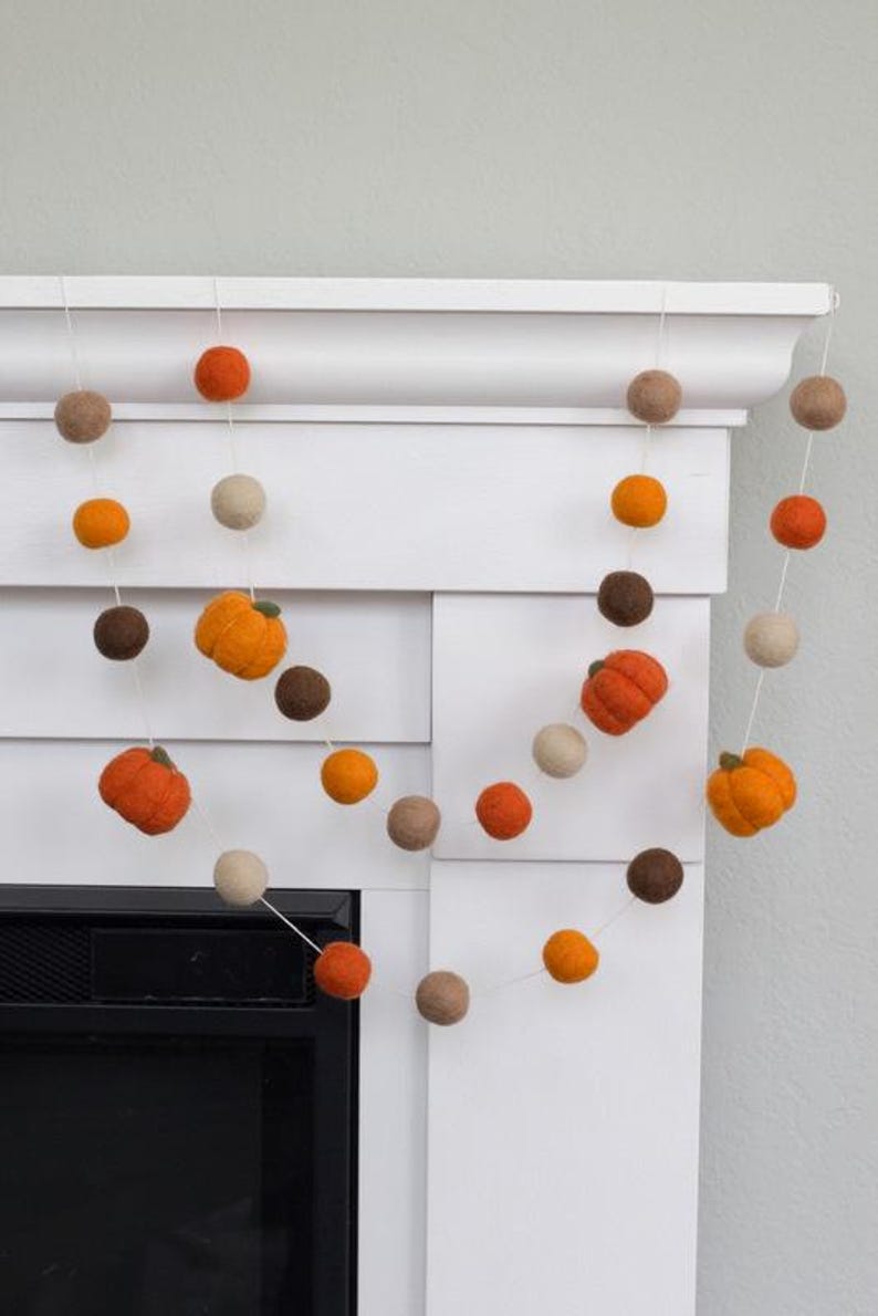 May include: A decorative garland featuring felted wool balls in shades of orange, brown, and cream, interspersed with small, textured felt pumpkins. The individual elements are strung together on a thin white cord, creating a repeating pattern.