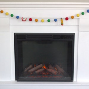 May include: A white fireplace mantel with a garland of colorful felt balls and felt cutouts of an apple, a pencil, and a musical note. The fireplace has a black metal surround and a realistic-looking electric fire.