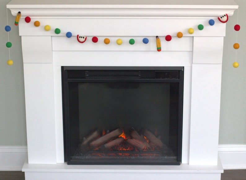 May include: A white fireplace mantel with a garland of colorful felt balls and felt cutouts of an apple, a pencil, and a musical note. The fireplace has a black metal surround and a realistic-looking electric fire.