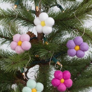 May include: Five colorful felt flower ornaments with yellow centers and silver wire hangers are displayed on a Christmas tree. The flowers are in shades of white, pink, purple, green, and magenta.