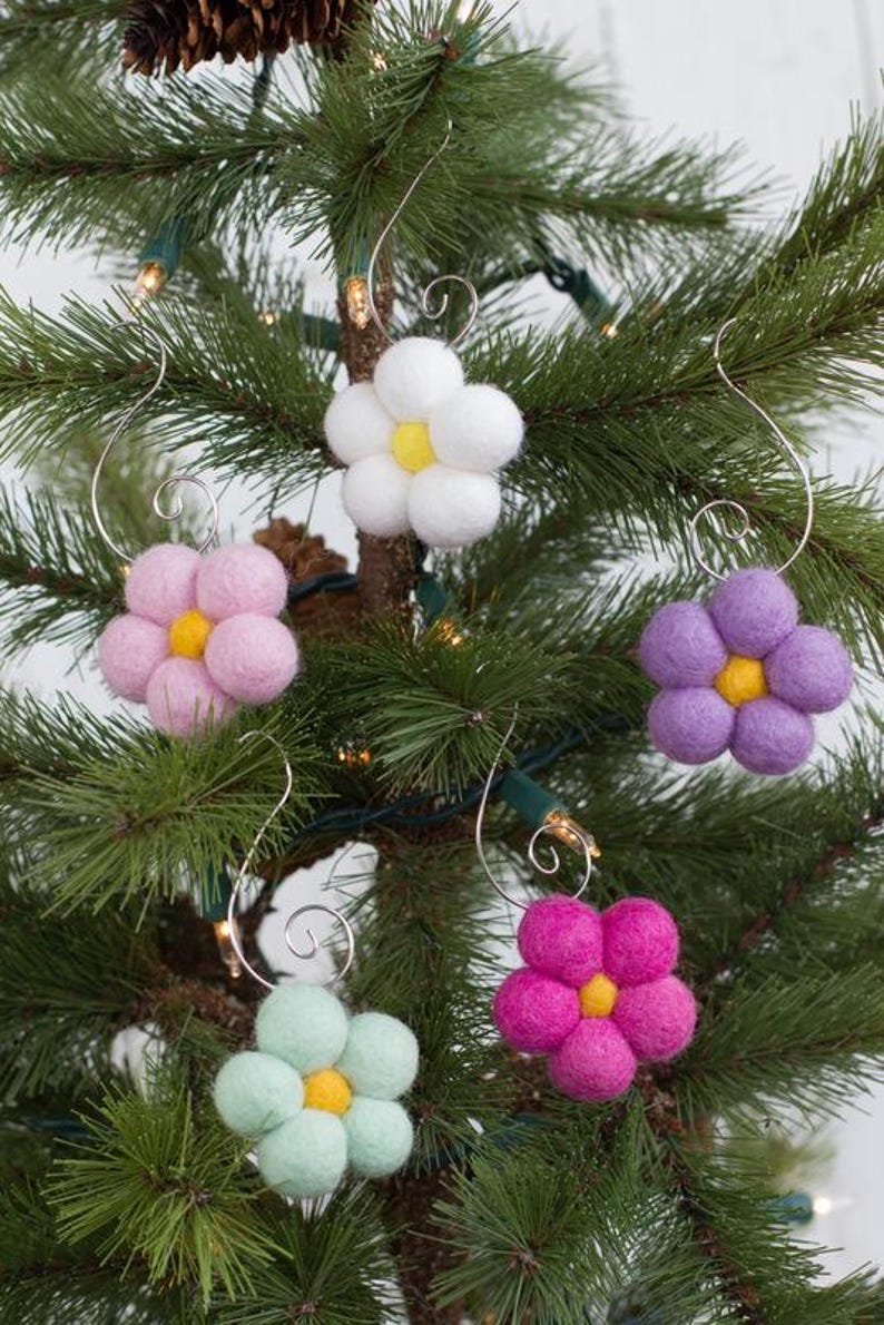 May include: Five colorful felt flower ornaments with yellow centers and silver wire hangers are displayed on a Christmas tree. The flowers are in shades of white, pink, purple, green, and magenta.