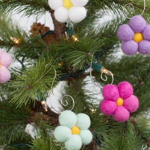 May include: A close-up of a Christmas tree decorated with handmade felt flower ornaments. The ornaments are in various colors, including pink, white, purple, green, and fuchsia, each with a yellow center. The ornaments are attached with silver wire.