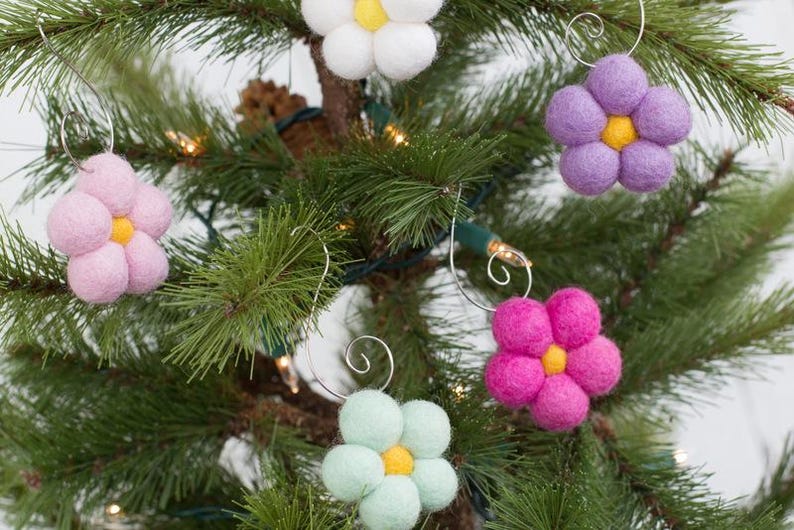 May include: A close-up of a Christmas tree decorated with handmade felt flower ornaments. The ornaments are in various colors, including pink, white, purple, green, and fuchsia, each with a yellow center. The ornaments are attached with silver wire.