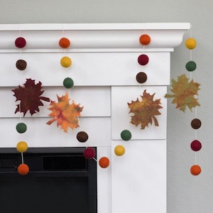 May include: A decorative garland featuring felted wool balls in shades of burgundy, orange, yellow, and dark green, interspersed with flat, die-cut felt maple leaves in variegated autumn colors, all strung together on a thin white cord.