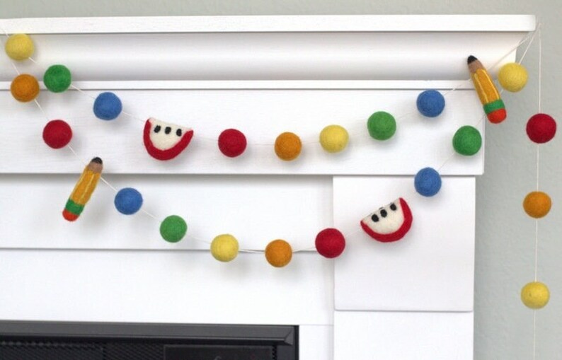 May include: A felt garland with red, yellow, blue, and green pom-poms, alternating with felt watermelon slices and a pencil. The garland is strung on a white background.