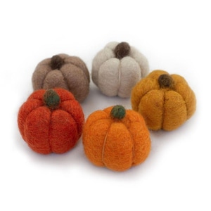 May include: Five felt pumpkins in shades of orange, brown, and cream are arranged on a white surface. Each pumpkin has a small, dark brown or green stem. The pumpkins vary in size and shape, creating a rustic autumn theme.