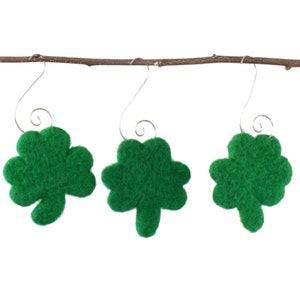 May include: Three green felt shamrock ornaments hanging from a brown twig. Each shamrock has a silver wire hook for hanging. The shamrocks are a vibrant green color and have a textured appearance.