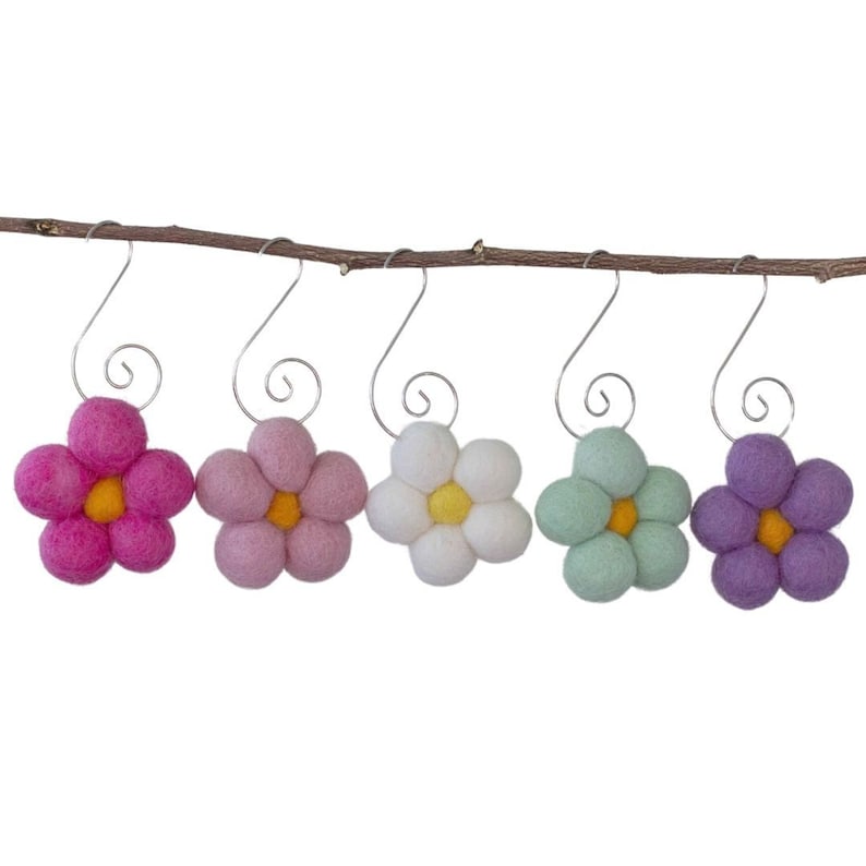 May include: Five felt flower ornaments in shades of pink, lavender, white, and green, each with a yellow center. They hang from silver hooks on a brown twig, creating a cheerful, spring-like display.