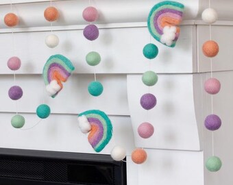 May include: A decorative garland featuring felted wool balls in shades of orange, pink, purple, mint green, and white, interspersed with three felted rainbow shapes that include small white cloud accents, all strung together on a thin white cord.