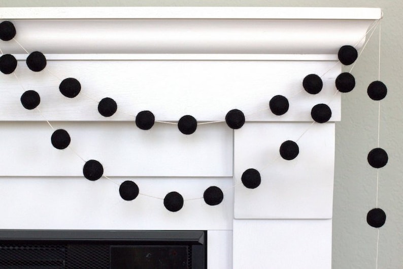 May include: A decorative garland consisting of numerous solid black felt balls strung together on a thin white cord. The spheres are evenly spaced along the string, creating a repeating pattern of dark circular shapes against the white background.