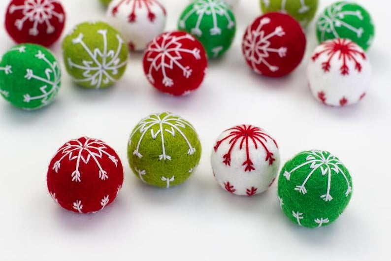 May include: Close-up view of numerous felt Christmas ornaments.  The ornaments are small, spherical, and feature embroidered snowflake designs. Colors include red, green, and white felt with white embroidery. The ornaments are arranged on a white surface, showcasing their festive design and handcrafted nature.  These are perfect for Christmas tree decorations or festive home decor.