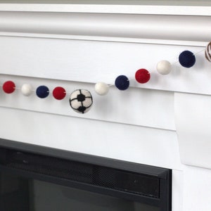 May include: A white, red and blue felt ball garland with a basketball, football and soccer ball hanging from it. The garland is hanging above a fireplace.