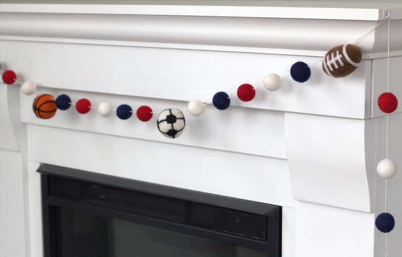 May include: A white, red and blue felt ball garland with a basketball, football and soccer ball hanging from it. The garland is hanging above a fireplace.