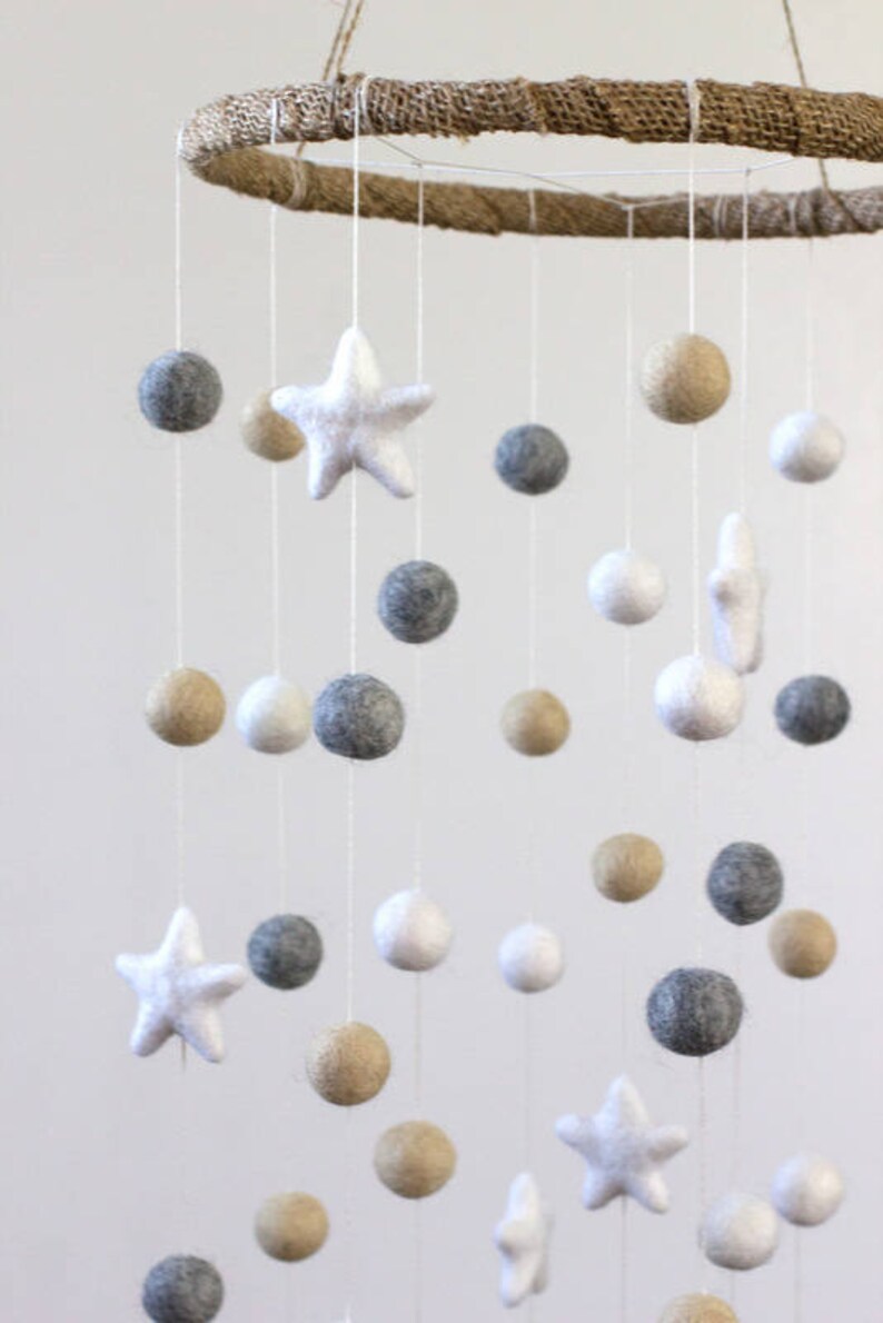 May include: A white, gray, and beige felt baby mobile with stars and balls hanging from a natural jute ring.