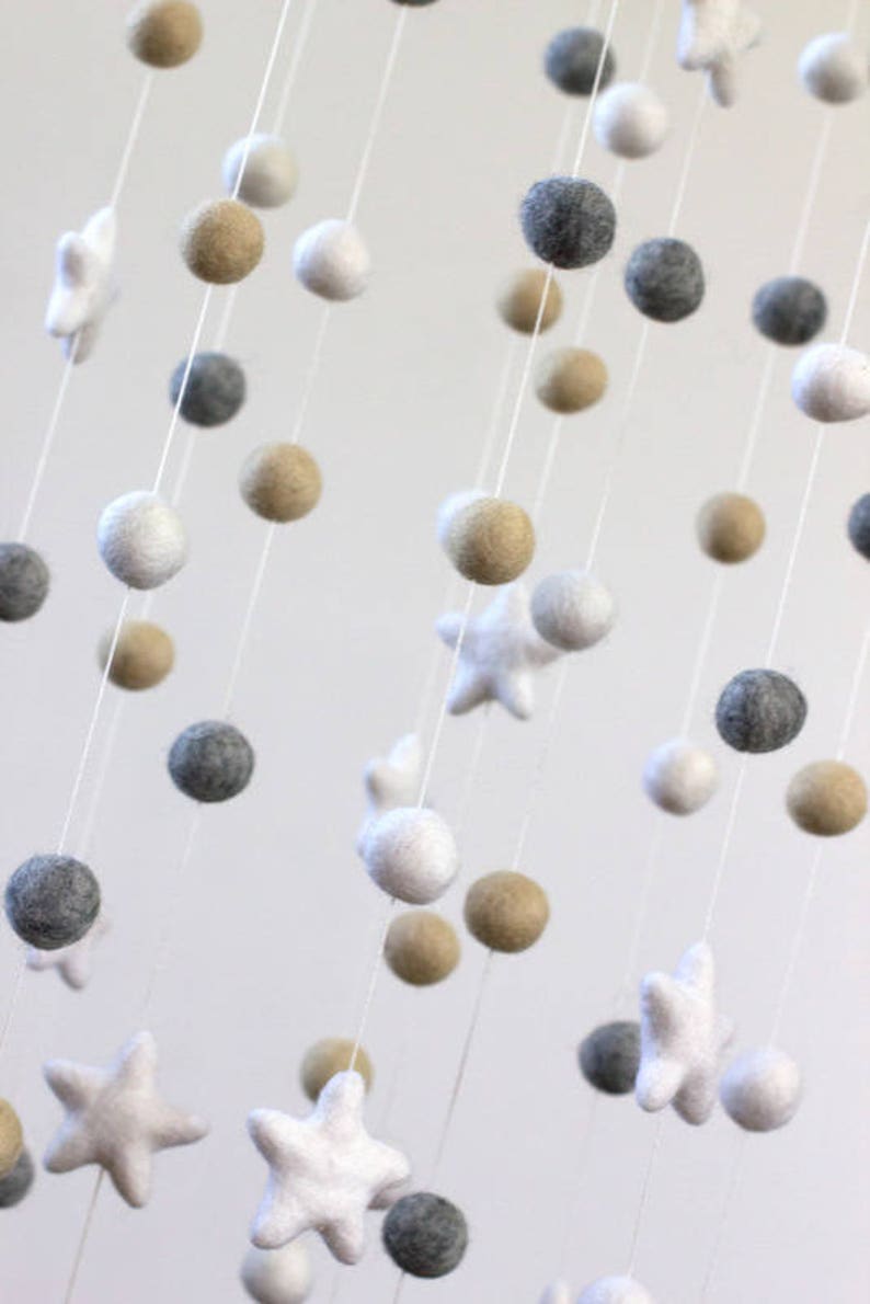 May include: A white, gray, and beige felt baby mobile with stars and pom-poms hanging from a white string.