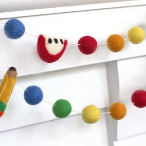 May include: A felt garland with colorful balls and felt cutouts of a pencil and fruit. The garland is strung on a white string and is hanging on a white wall.