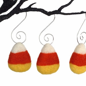 May include: Three felt candy corn ornaments hanging from a black branch. The ornaments are white, orange, and yellow.