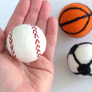May include: Three felt balls representing different sports: a white baseball with red stitching, an orange basketball with black lines, and a white football with black panels.