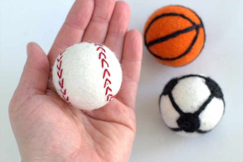 May include: Three felt balls representing different sports: a white baseball with red stitching, an orange basketball with black lines, and a white football with black panels.