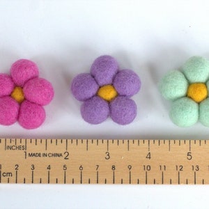 May include: Three felt flower decorations in pink, purple, and mint green, each with a yellow center. A wooden ruler with inch and centimeter markings is placed below the flowers. The flowers are approximately 2 inches in diameter.