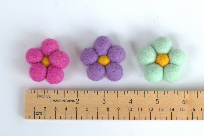 May include: Three felt flower decorations in pink, purple, and mint green, each with a yellow center. A wooden ruler with inch and centimeter markings is placed below the flowers. The flowers are approximately 2 inches in diameter.