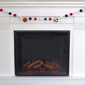 May include: A white fireplace mantel with a garland of white, red and blue pom-poms and felt sports balls. The fireplace has a black electric insert with a realistic flame effect.