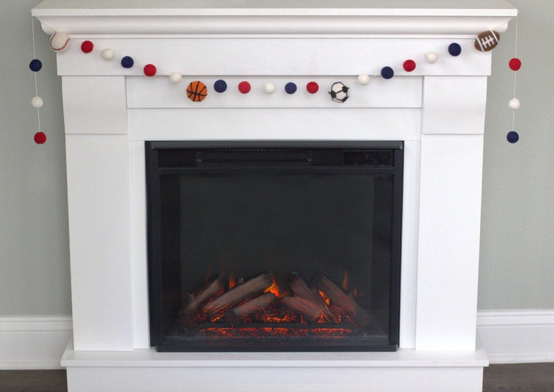 May include: A white fireplace mantel with a garland of white, red and blue pom-poms and felt sports balls. The fireplace has a black electric insert with a realistic flame effect.