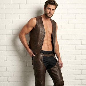 May include: A dark brown leather vest featuring a button-front closure and a pair of matching dark brown leather chaps worn over dark trousers. The vest has a tailored fit and the chaps are secured with a belt at the waist, creating a coordinated set.