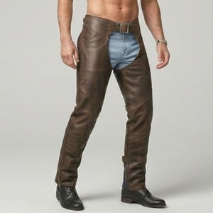 Puede incluir: Chaps de cuero marrón oscuro con un cinturón en la cintura con hebilla de metal, paneles laterales en las piernas y un acabado liso. Los chaps se usan sobre pantalones vaqueros azul claro, extendiéndose desde la cintura hasta los tobillos.