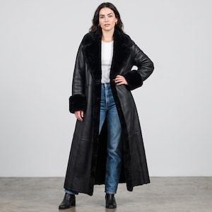 Könnte beinhalten: Ein langer schwarzer Ledermantel mit einem breiten schwarzen Kunstfellkragen und passenden Fellmanschetten an den Ärmeln. Der Mantel wird über einem weißen Oberteil mit Rundhalsausschnitt und blauen Jeans getragen, kombiniert mit schwarzen Lederstiefeletten.