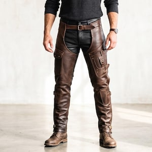 May include: Dark brown leather chaps featuring a waist belt with a metal buckle, multiple side pockets with flap closures, and adjustable straps with buckles along the legs. The chaps are worn over black pants and paired with brown leather boots.