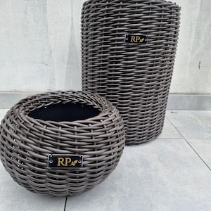 May include: Two dark brown woven planters with a rounded, textured exterior. Each planter features a small, rectangular black plaque on the front with the gold letters RP and a small leaf icon. The planters have a hollow, open top design.