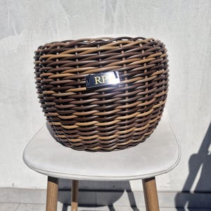 Puede incluir: Una cesta redonda tejida con hebras de mimbre sintético que alternan entre marrón oscuro y bronceado claro. Una pequeña placa rectangular negra está unida al frente, con las letras RP en color dorado en una fuente con serifa.