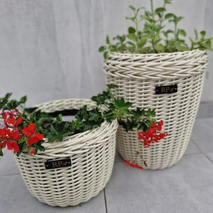 May include: Two off-white woven planters with a round shape and a braided rim. Each planter features a small black rectangular tag with the letters RP and a leaf icon. The shorter planter holds red geraniums, while the taller one holds green foliage.