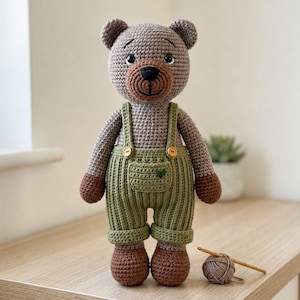 Peut inclure: Un ours en peluche crocheté avec une fourrure marron clair, un museau marron foncé et des yeux de sécurité noirs. L'ours porte une salopette vert olive avec deux boutons en bois sur les bretelles et une petite poche avant avec un cœur vert foncé.