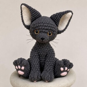 Peut inclure: Une figurine de chat crochetée gris foncé avec de grandes oreilles pointues dotées de sections intérieures couleur crème. Le chat a des yeux ambrés, de fines moustaches noires et de petits coussinets rose clair sous les pattes.