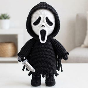 Peut inclure: Une poupée crochetée à la main représentant un masque blanc avec des découpes noires pour les yeux et la bouche, vêtue d'une robe à capuche noire aux bords effilochés. La figurine tient un petit couteau crocheté gris clair à la main.