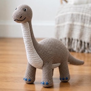 Puede incluir: Un juguete de dinosaurio tejido a ganchillo en color marrón claro con un panel de color crema en el pecho y el cuello. El juguete presenta ojos de plástico negro, una boca cosida y pequeños detalles bordados de estrellas azules en los pies.