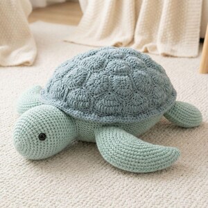 Peut inclure: Une peluche tortue au crochet avec un corps vert menthe clair et une carapace texturée vert sauge plus foncé. La tortue présente une tête arrondie avec un œil en plastique noir visible et quatre nageoires s'étendant du corps principal.