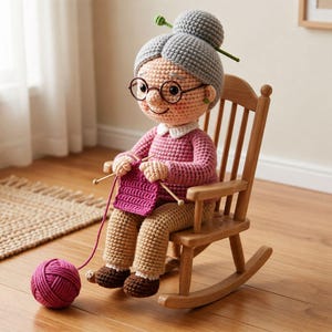 Peut inclure: Une poupée crochetée représentant une personne âgée aux cheveux gris en chignon, portant des lunettes rondes, un pull rose à manches longues et un pantalon beige. La poupée est assise dans une petite chaise à bascule en bois avec du tricot.