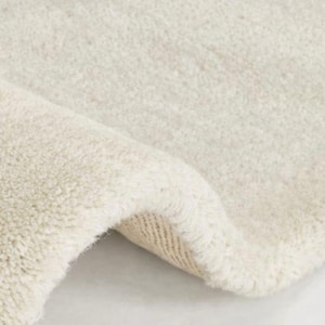 May include: A plush, off-white area rug with a thick, soft pile texture. The rug is folded over at one corner, revealing the woven backing material underneath the dense, light-coloured fibres that cover the entire surface of the floor covering.