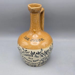 May include: A vintage ceramic whiskey jug with a tan glazed neck and a cream-colored base. The body features black floral illustrations and text reading THE JORUM, RODERICK DHU OLD HIGHLAND WHISKY, WRIGHT & GREIG LTD, GLASGOW. It has a single side handle.