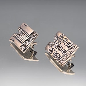 May include: A pair of silver-toned metal clip-on earrings with a rectangular, slightly curved shape. Each earring features an embossed pattern of geometric lines, small circles, and floral-like motifs across the front surface.
