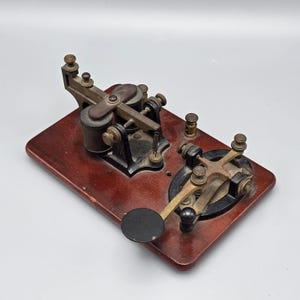 May include: A vintage telegraph key mounted on a rectangular dark wood base. The device features black metal components, brass adjustment screws, a circular black knob, and an electromagnetic coil assembly with exposed wiring and metal springs.