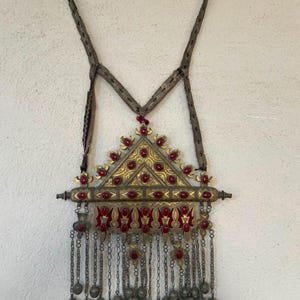Puede incluir: Un gran colgante triangular de metal con intrincados grabados en tono dorado y piedras ovaladas rojas incrustadas. La pieza incluye una barra horizontal, una sección inferior con detalles en esmalte rojo y múltiples cadenas colgantes con pequeñas campanas.